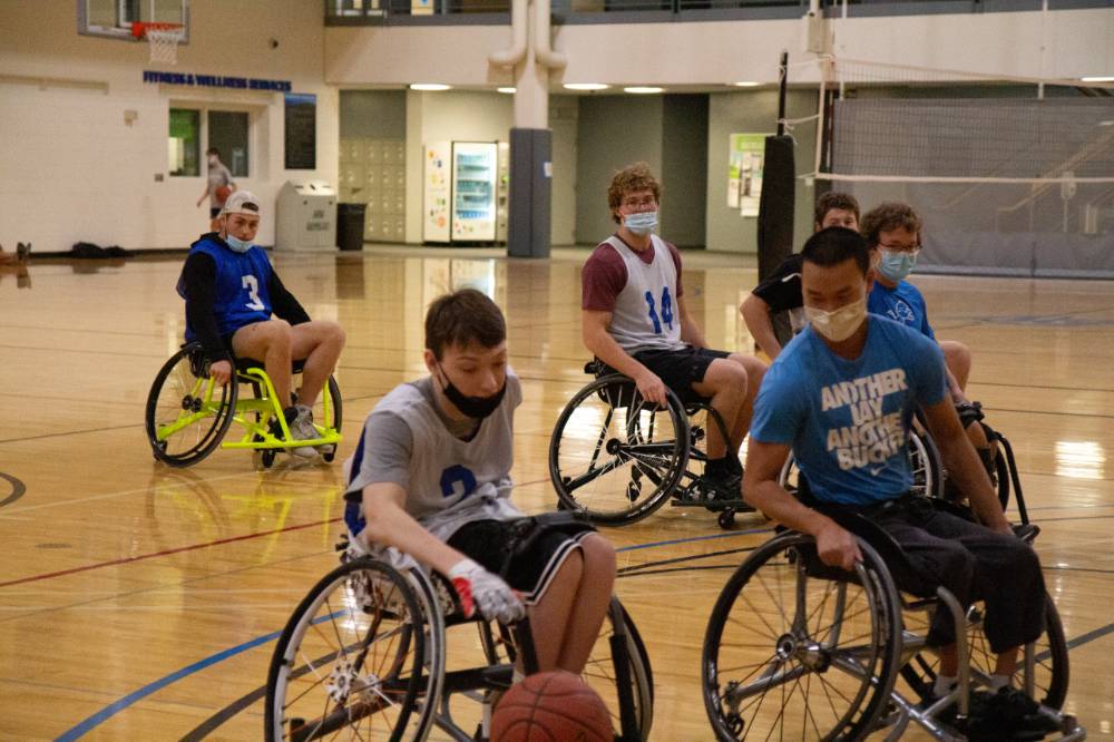 Adult dribbling a basketball in a wheelchair with other participants defending the ball.